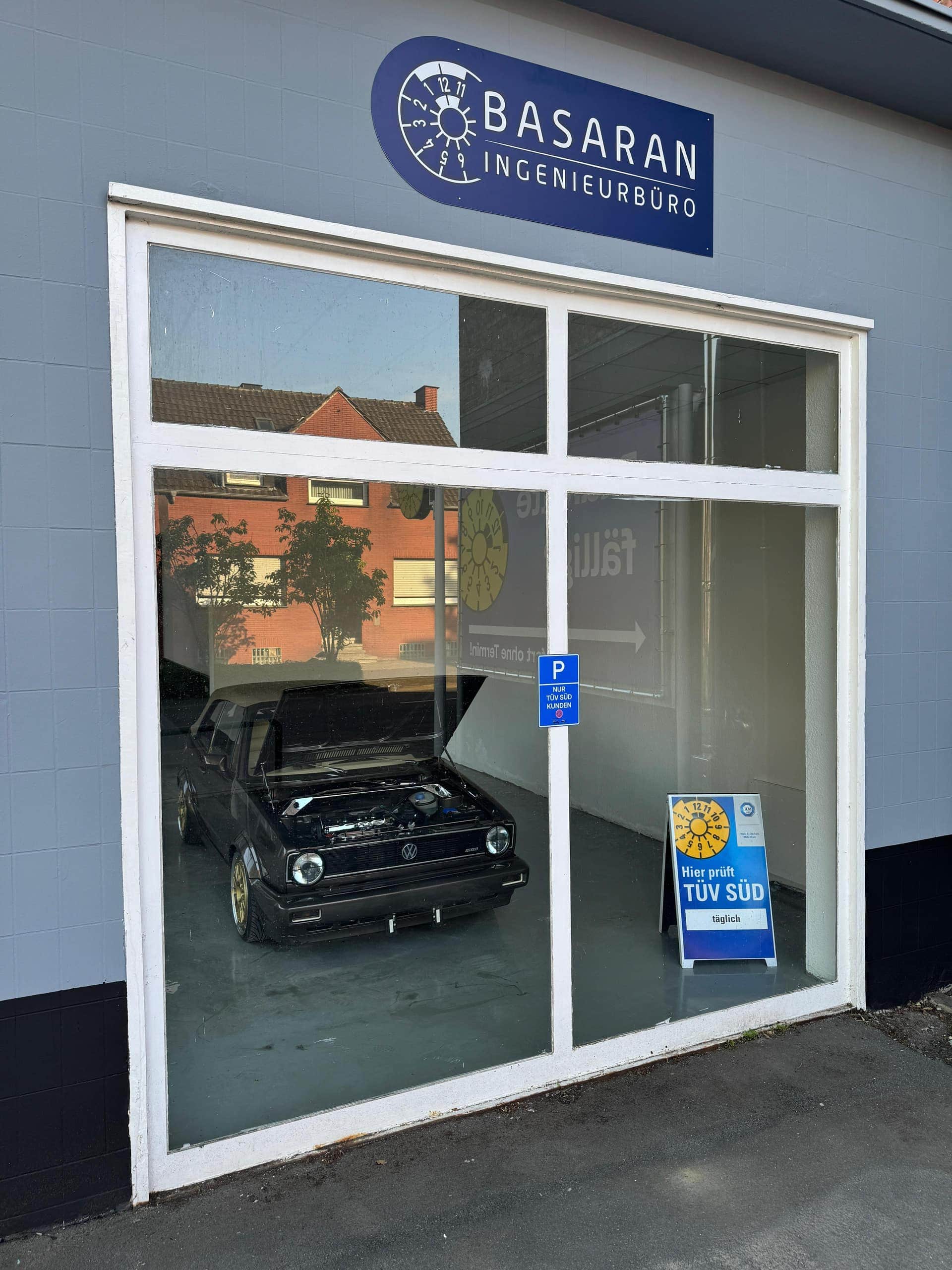 Blick durch das Schaufenster der Prüfhalle auf einen schwarzen VW Golf Cabrio mit geöffneter Motorhaube, darüber das Schild Basaran Ingenieurbüro